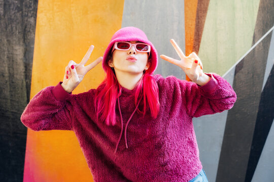 Emotional young hipster woman with pink hair and sunglasses in magenta fluffy sweatshirt and bucket hat making V sign by fingers on graffiti background. Urban street fashion. Gen Z self expression.