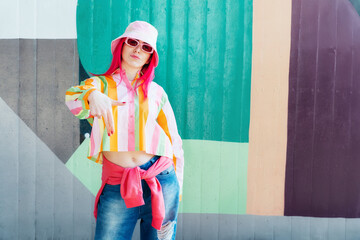 Young woman with pink hair and sunglasses in Bucket hat and multicolor strippled shirt posing on graffiti wall background. Street artist with mural, hipster, blogger. Urban street fashion - obrazy, fototapety, plakaty