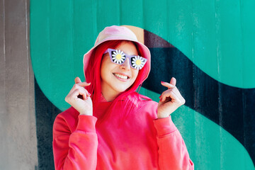 Vanilla Girl. Kawaii vibes. Candy colors design. Young woman with pink hair and sunglasses with flower stickers in bucket hat making making Finger Heart sign on graffiti wall background.. Love fashion - Powered by Adobe