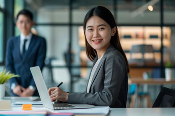 Smiling Young Businesswoman Working on Laptop With Colleague in Background During Office Hours