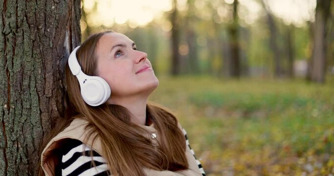 Woman leaning against a tree listening to music on white headphones