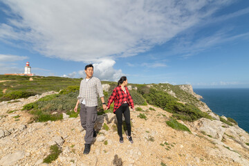 A couple is walking on a rocky hillside with a lighthouse in the background