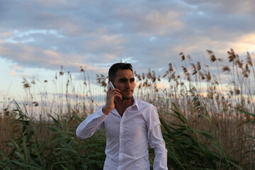 A man in a white shirt stands in a field of tall grass. The sky is cloudy and the sun is setting