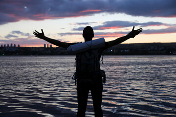 A traveler man stands on the beach with a backpack. He throws his arms up in the air as if he's...