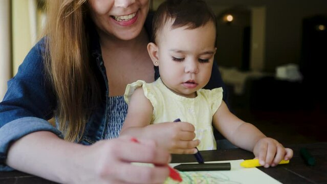 Latin mother having fun with baby girl painting with colors at home. Latin family, mom and daughter relationship concept