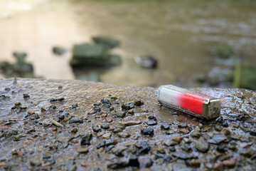 Discarded single use electronic cigarette vape left on concrete wall edge beside a murky stream with rocks littered around. - obrazy, fototapety, plakaty