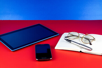 Tablet Device with Mobile Phone Notebook Pen and Spectacles on a Red Surface and Blue Background - 785706254