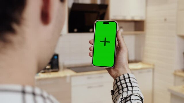 Close-up of the young man in casual clothes, holding a smartphone with a mockup green screen, back view, with the background of cozy home kitchen