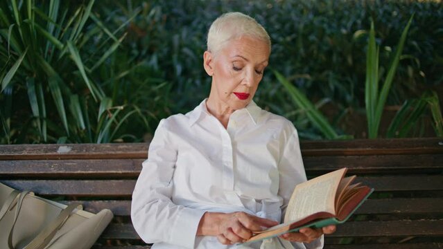 Grey hair lady reading book in city park. Focused senior flipping novel pages