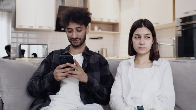Young offended upset woman sitting on the couch at home next to a young man with a smartphone who indifferent of her problems. Concept of difficulties in a couple relationship