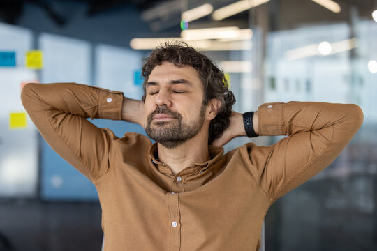 A middle-aged man with curly hair relaxes by stretching his arms behind his head in a contemporary office atmosphere, demonstrating a moment of stress relief.