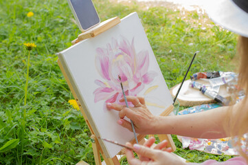 Pink lotus painting on canvas standing on easel outdoor. Woman artist drawing in summer part on a...