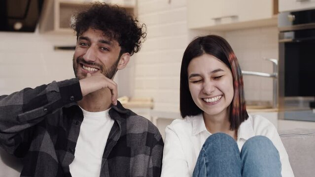 Young cheerful couple in casual clothes sitting on a couch in a kitchen and laughing loud when looking at the camera. Concept of happiness and joy in the relationship