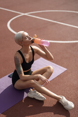 Fitness enthusiast taking a break to drink water on a sunny day at the athletic track with yoga mat.
