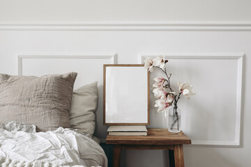 Blank wooden picture frame mockup on old book. Wooden night stand with fluted glass vase. Blooming magnolia tree branches. Scandinavian interior. Elegant bedroom. White wall background, stucco decor.