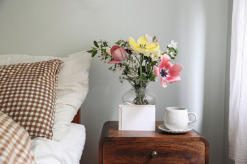 Spring interior still life. Blank greeting card, invitation mockup. Elegant bedroom. Tulips, cherry tree blossoms bouquet in glass vase. Wooden night stand. Cup of coffee. Checkered bedding, cushions.