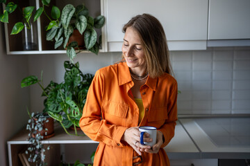 Pleased woman enjoy morning routine with coffee at home rest in kitchen surround by houseplants...