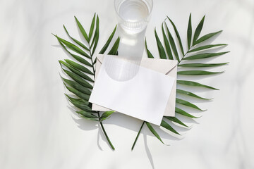 Blank paper, greeting card. Invitation mockup with fresh green palm leaves. Drinking glass, water in sunlight. Shadows on white table background. Flat lay, top view. Aesthetic business brand template