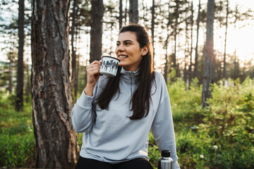 Young caucasian woman pouring and drinking water in forest while taking a break from hiking
