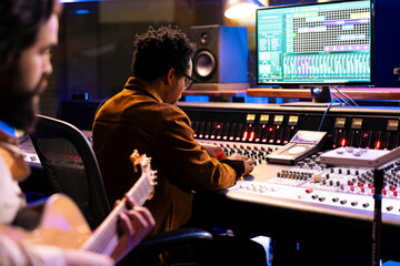 Skilled songwriter artist recording a song with electro acoustic guitar tunes, making music for his pop rock album. Musician singing tracks on instrument in control room, works with audio expert.