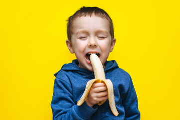 A small funny cheerful ukrainian boy bites a banana, makes faces and plays around. Studio photography with yellow background. - obrazy, fototapety, plakaty