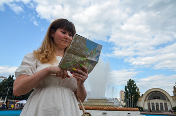 Portrait of pretty female traveler on the city square holding a map in her hands with blue sky with clouds on background, concept of summer holidays and tourism