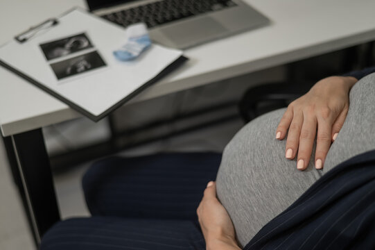 A pregnant woman works on a laptop in the office and looks at a photo from an ultrasound scan of the fetus. Belly close-up.