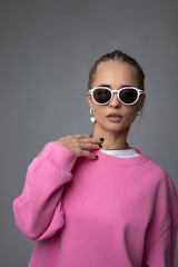 Beautiful blonde woman in sunglasses, pink hoodie and white panties posing on gray background
