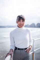 Young woman, white sweater, brown hair, smiles by Gijón's seaside, enjoying coastal tranquility.