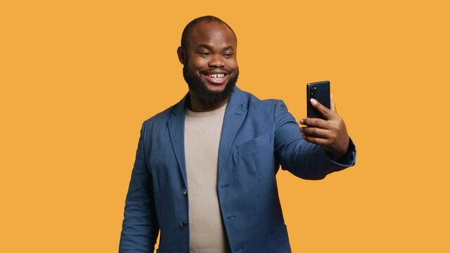 Joyous man using smartphone to take selfies and post them on social media. Happy BIPOC person taking photos using phone selfie camera, isolated over yellow studio background, camera A