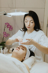 A girl cosmetologist spreads facial foam for a patient on a massage table.