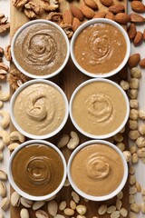 Many tasty nut butters in bowls and nuts on white table, flat lay