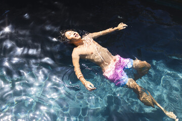 A teenage Asian boy is floating in a pool outside at home, wearing a pink swim bottom, copy space