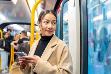 Asian woman using mobile phone and listening to the music on earphones during travel on train in the city. Attractive girl enjoy urban lifestyle in the city with using wireless technology on device.
