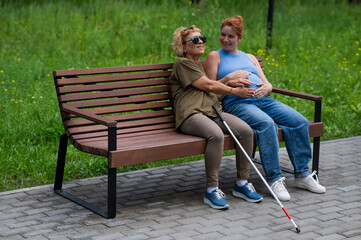 An elderly blind woman holds her pregnant daughter by the belly while sitting on a bench in the...