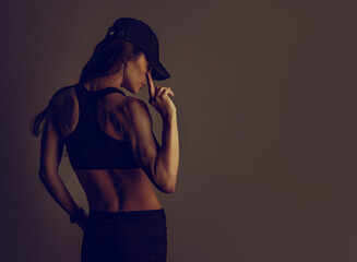 Fuck you. Woman in black sport bra and summer black cap showing the fuck sign the hand, standing back side on dark shadow studio background. Body closeup toned color banner - obrazy, fototapety, plakaty