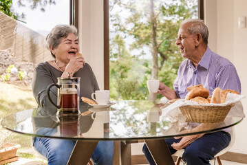 An older Latino man and woman sitting in the dining room of their home chatting and drinking coffee.