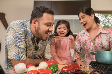 Daughter giving her father, in a wooden spoon, a sample of sauce that she has cooked with her mother.
