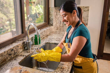 A beautiful Latin woman with a yellow apron and gloves washes the dishes in the kitchen.