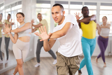 Fototapeta na wymiar Expressive young Asian male instructor in white t-shirt and cargo pants leading modern dance exercise class for multiethnic group in well-lit fitness studio..