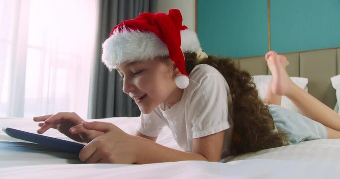 Kid in Christmas hat Santa Clausa watching online video under covers with digital tablet network. Child on social media. Child with digital tablet. Kid dream online video games at Christmas.