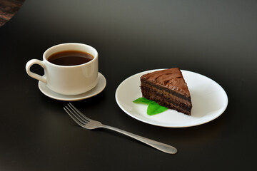 A piece of chocolate layer cake with mint leaves, a fork, and a cup of black coffee on a saucer on a black background.