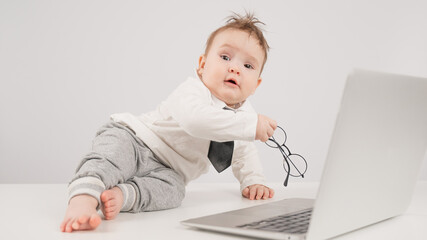 A cute child wearing a suit holds glasses and lies behind a laptop.