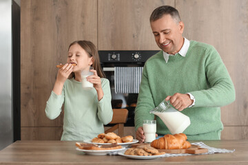 Little girl with her father eating different pastries and drinking milk at table in kitchen