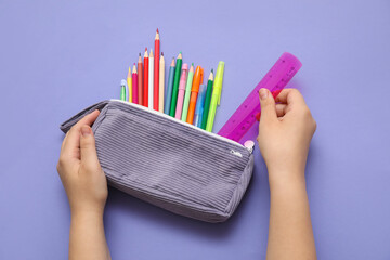 Child's hands with pencil case and different school stationery on purple background