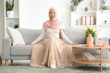 Beautiful young Asian woman in hijab sitting on sofa at living room