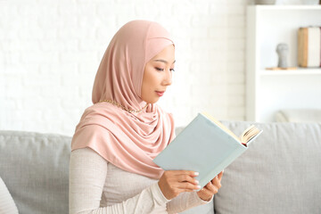 Beautiful young Asian woman in hijab sitting on sofa at living room and reading book