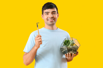 Young man with florarium and rake on yellow background