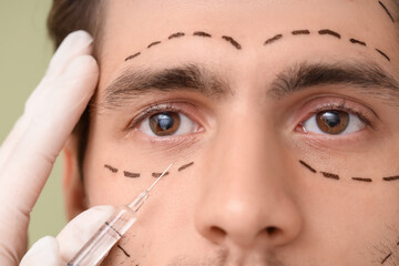 Young man with marked face receiving injection on green background, closeup. Plastic surgery concept
