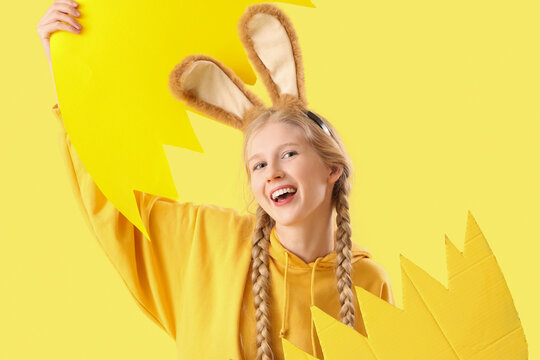 Young girl with Easter bunny ears and broken paper egg on yellow background